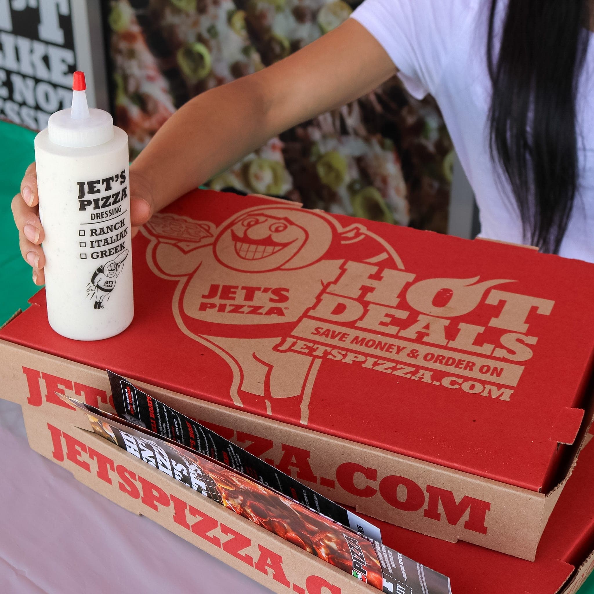 Hand holding a Jet's Pizza ranch dressing bottle over a stack of red Hot Deals pizza boxes