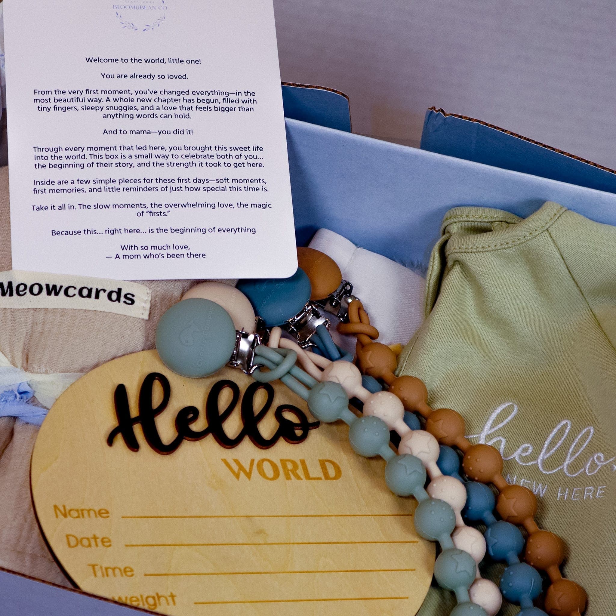 Open baby gift box with a welcome card, Hello World wooden sign, and pacifier clips in muted tones