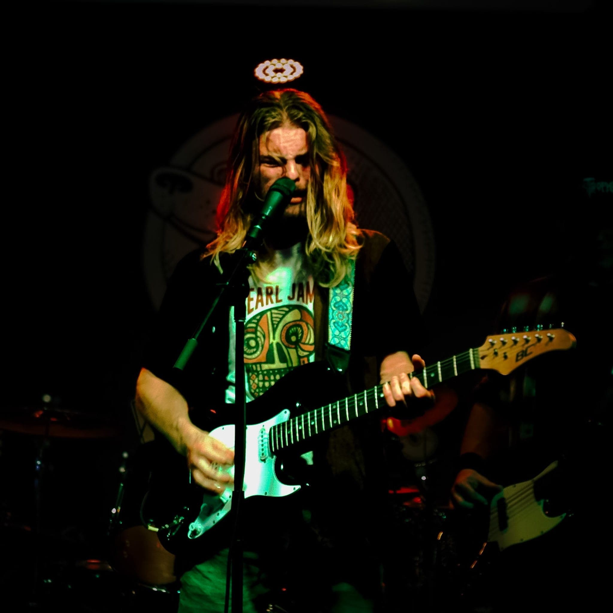 Rock vocalist playing guitar at the mic under green stage light, long hair rimmed red by the backlight