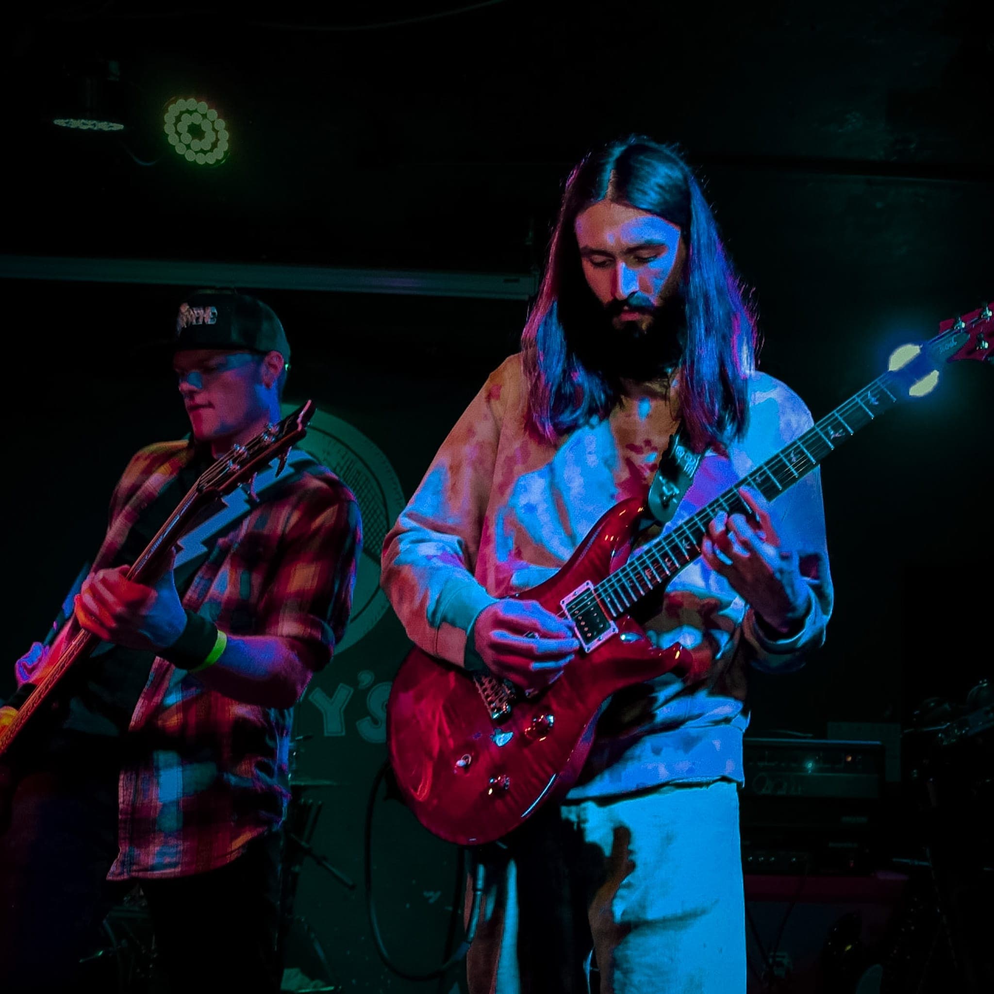 Two guitarists trading parts in blue and magenta light, one on a flame-red PRS