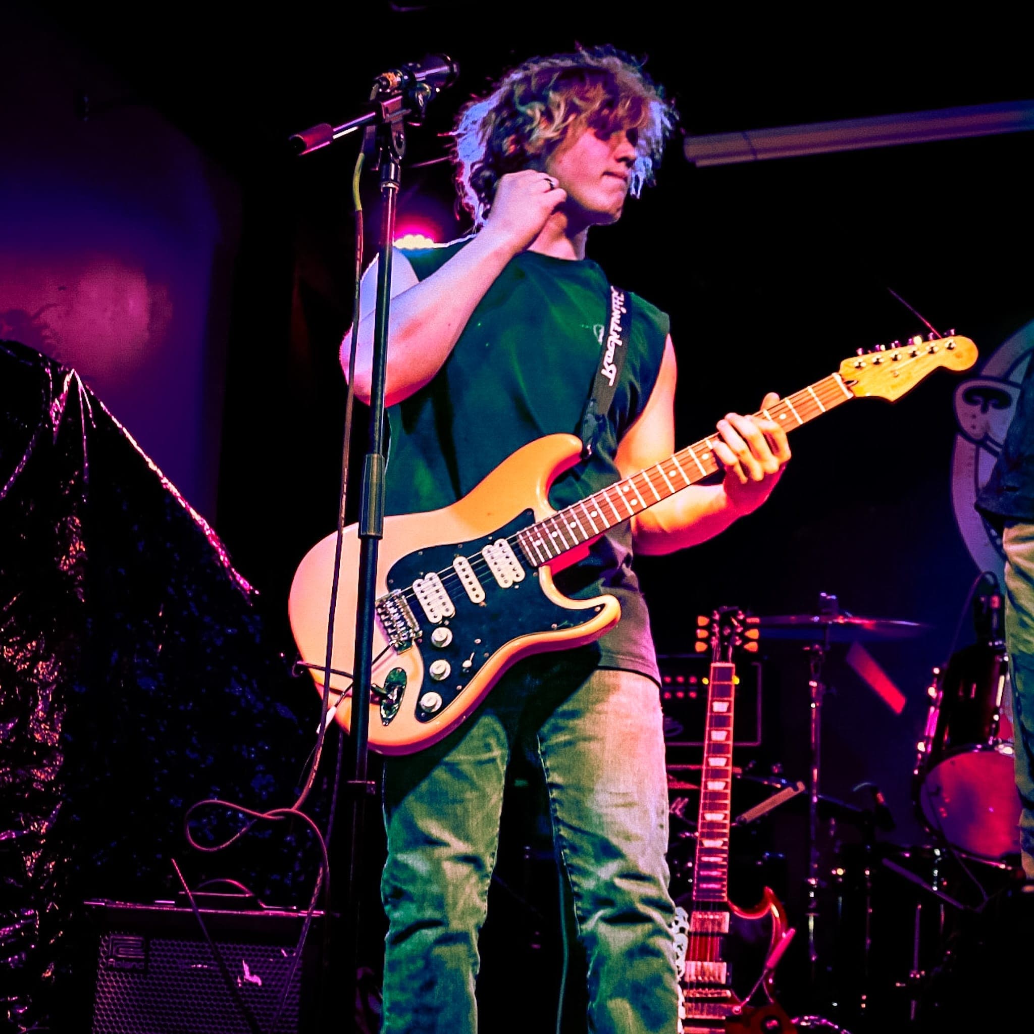 Guitarist in a sleeveless shirt playing a cream Stratocaster, washed in pink and violet stage light