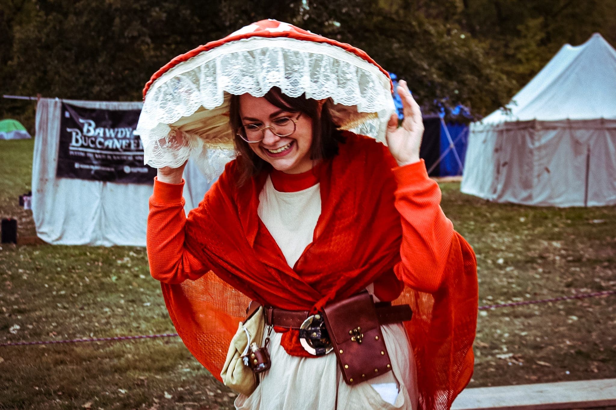 Festivalgoer laughing under a lace-trimmed mushroom-cap hat, tents behind her
