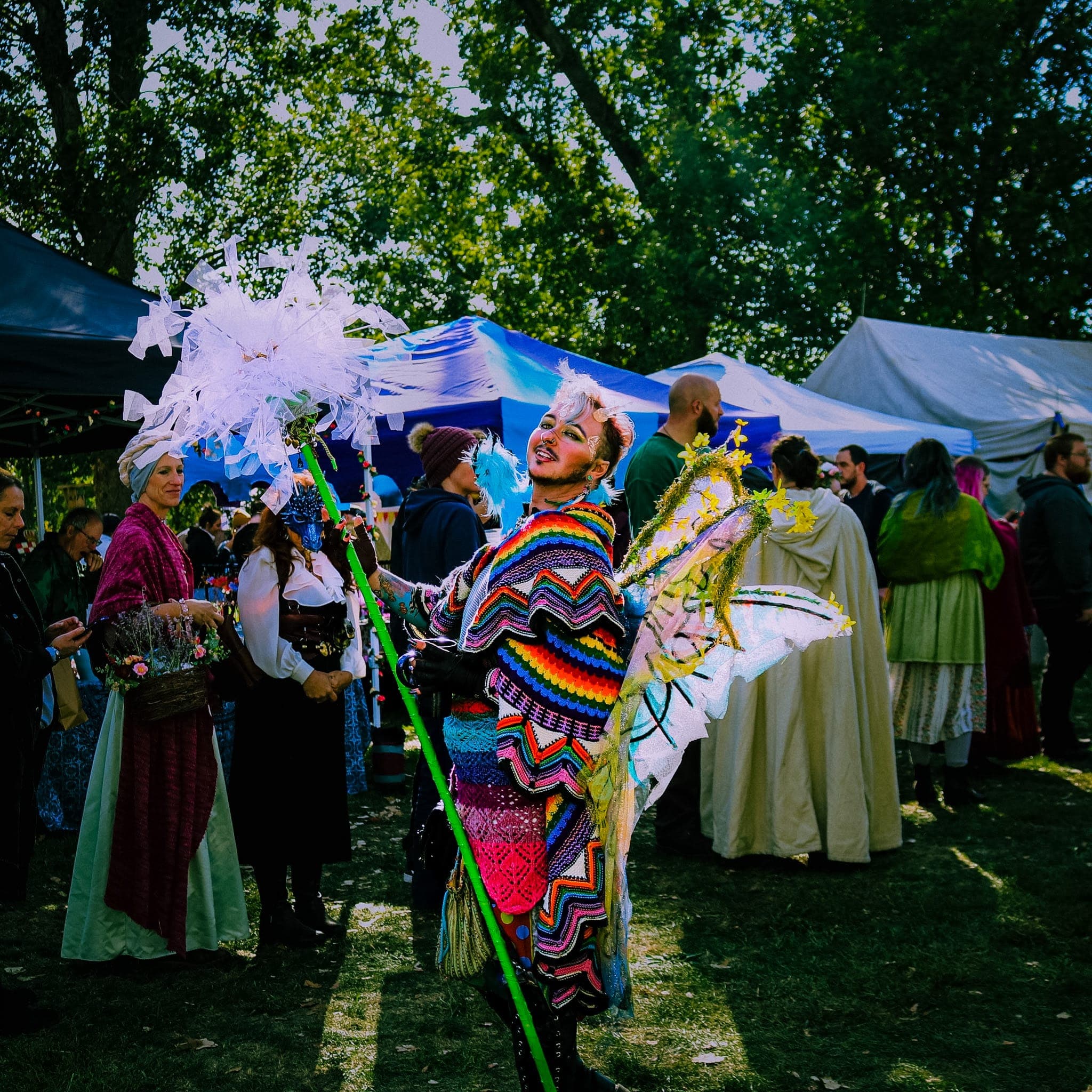 Fairy in glowing wings and a rainbow crochet wrap posing with a staff at a festival market