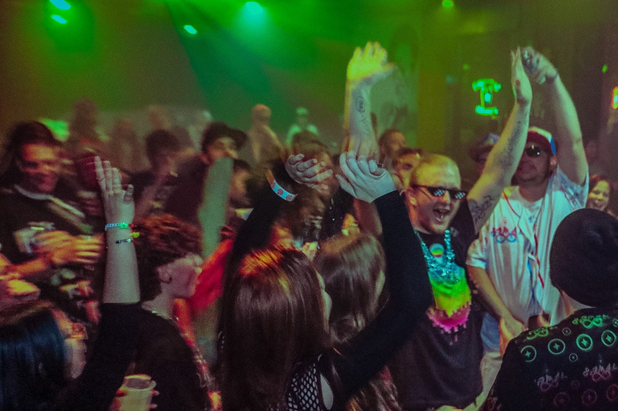 Crowd dancing under a green wash, hands in the air and faces mid-shout