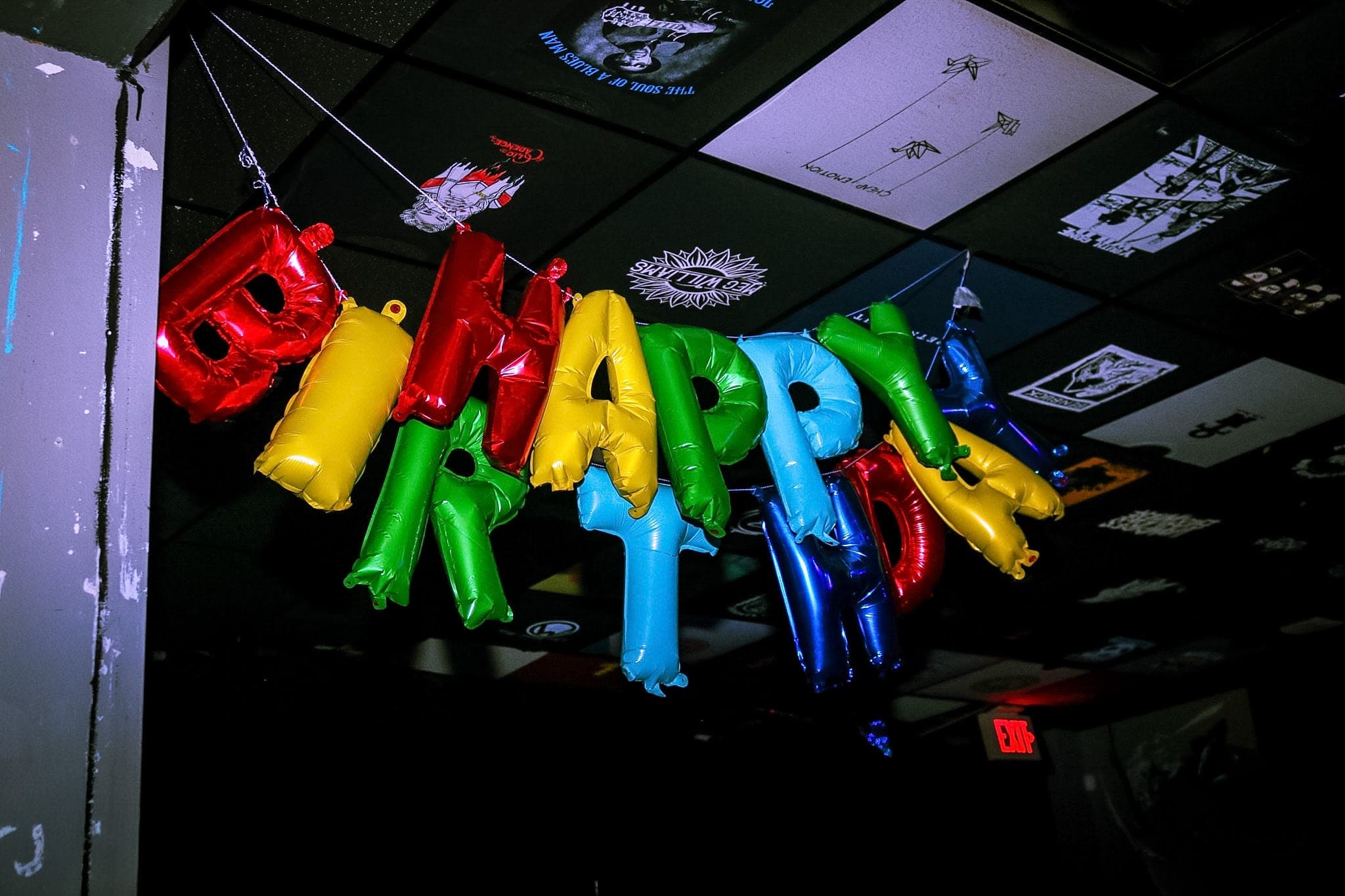 Happy Birthday balloon letters strung across a venue ceiling papered with gig posters